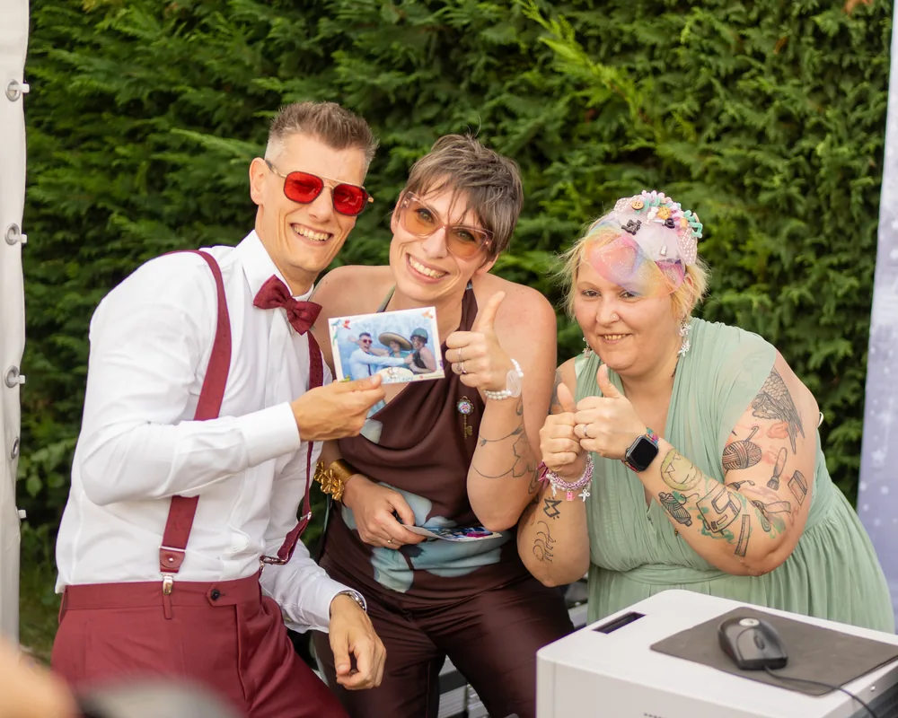Tre invitati sorridono verso l'obiettivo con i pollici in su; quello al centro tiene in mano la stampa 10x15 appena uscita, con la cornice chiara dell'evento. A destra, sul tavolo, la stampante della postazione.