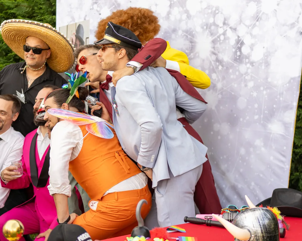 Un gruppo di invitati travestiti si accalca davanti al fondale: parrucca afro, sombrero e occhiali scuri, berretto da ufficiale, ali di fata sopra un gilet arancione e abiti fucsia e azzurri.