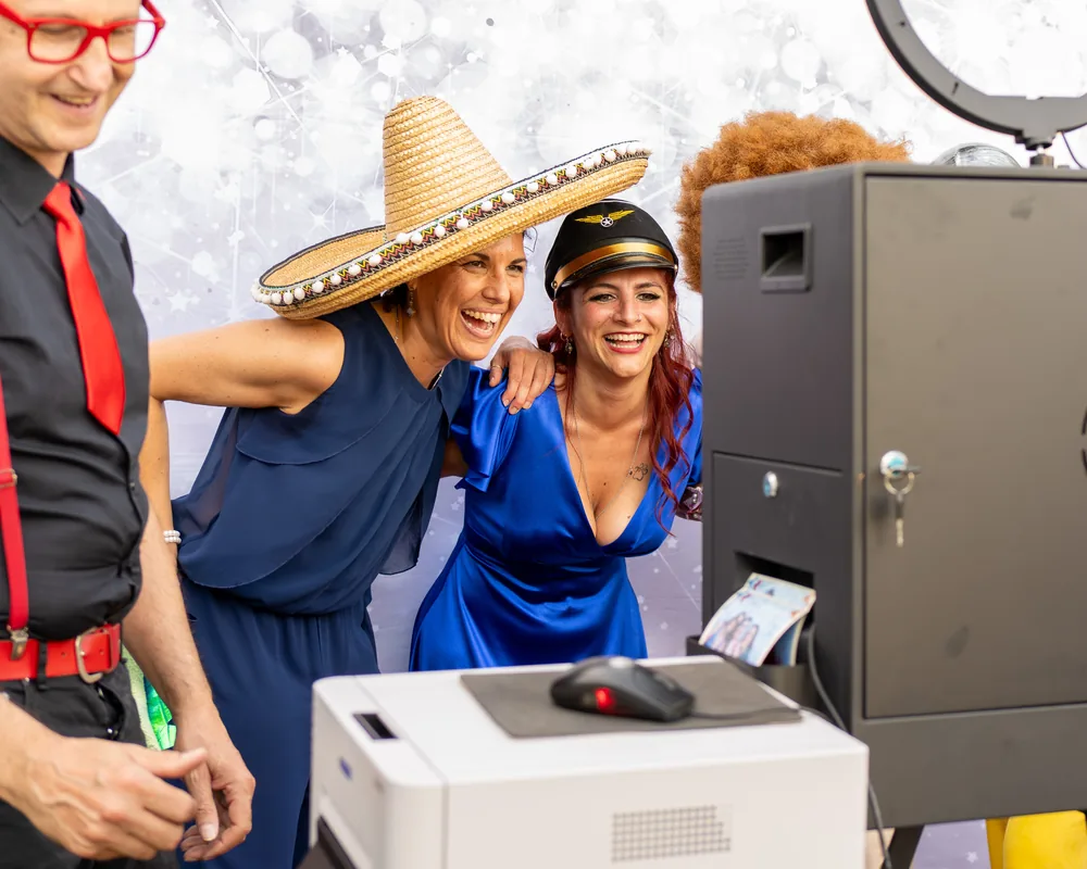 Due invitate, una con sombrero e una con cappello da aviatore, ridono davanti alla cabina del selfie; dalla fessura sotto lo sportello esce la stampa appena fatta e accanto si vede la stampante.