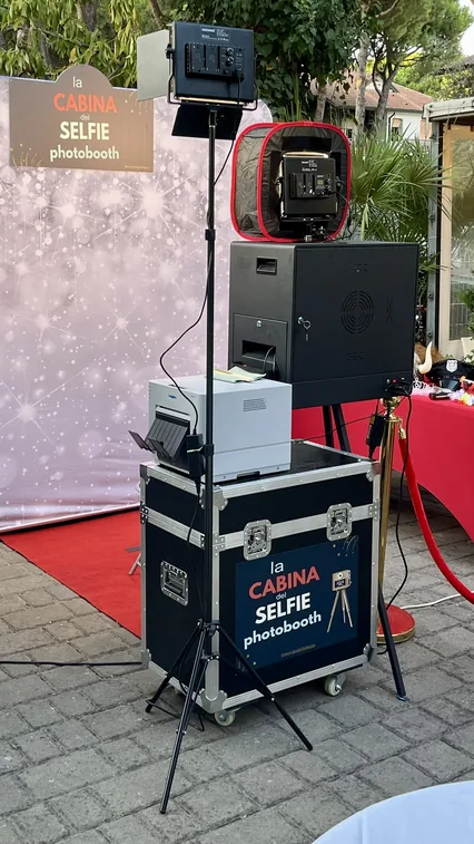 La postazione montata su un cortile: fondale con il cartello della cabina, luce LED, macchina fotografica, stampante sul flight case, tappeto rosso e cordone d'oro.