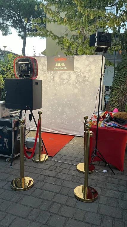 La postazione del photobooth montata in giardino al tramonto: colonna nera con fotocamera e diffusore rosso, fondale argentato con l'insegna, tappeto rosso, colonnine dorate con cordone e, a destra, il tavolo con gli accessori