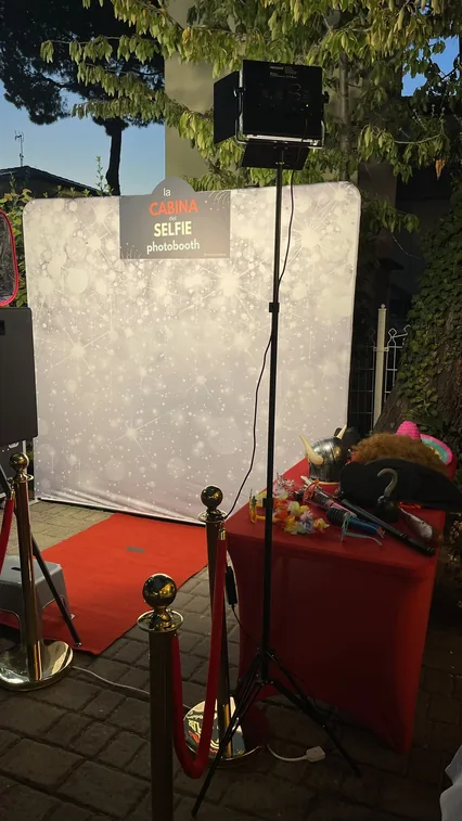 Il fondale argentato con l'insegna La Cabina del Selfie illuminato da un faretto su stativo, il tappeto rosso davanti e, a destra, il tavolo con parrucche, cappelli, occhiali e collane di fiori