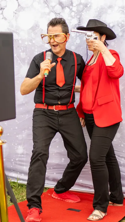 Un uomo con occhiali da Elvis canta al microfono accanto a una donna in rosso con cappello nero da cowboy, sul tappeto rosso davanti al fondale argentato della cabina.