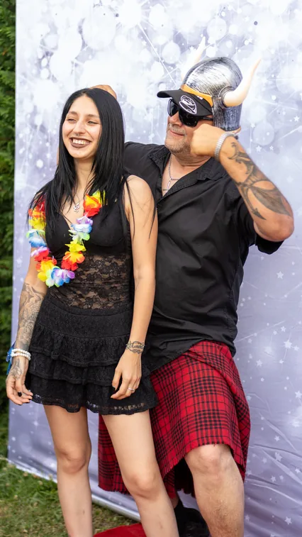 Una ragazza con collana di fiori colorati e un uomo con elmo vichingo, occhiali scuri e kilt a quadri rossi posano davanti al fondale argentato.
